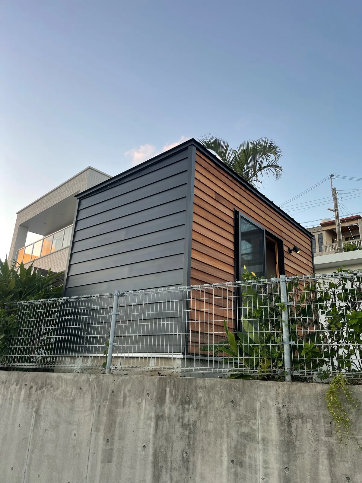 木の板とグレーの金属を組み合わせた外壁の小屋。背後に住宅と椰子の木